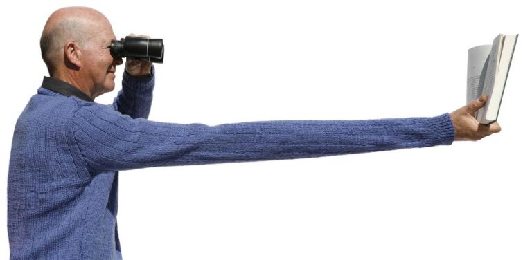 Middle age man in a blue jumper holding a book at arms length viewing through binoculars