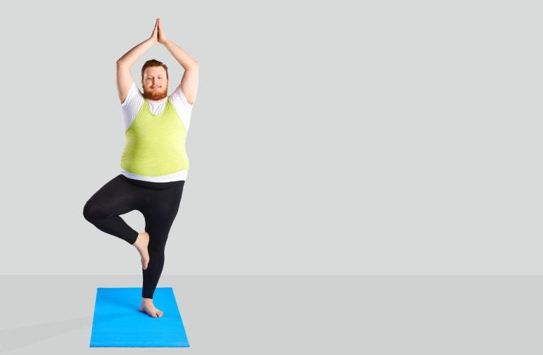 Fat man in tight yoga clothing standing on a blue mat on one leg with arms above head